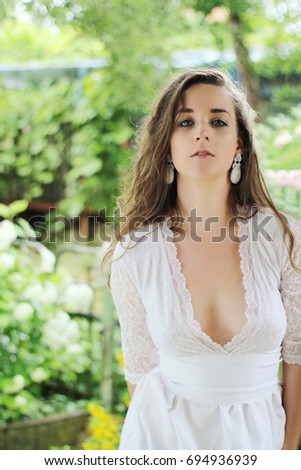 Young girl with brown hair down wearing a white wedding dress with deep neckline and white earrings. Picture taken outdoors in park after rain.