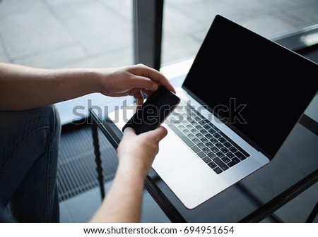 Businessman holding mobile phone with empty screen, sitting at glass table with open laptop computer with copy space on display.
Male using cell telephone and notebook for work.