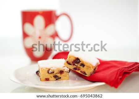 Two squares of freshly baked Sultana and Cranberry Slice on a white plate with a red napkin, a red mug and photographed on a white background.