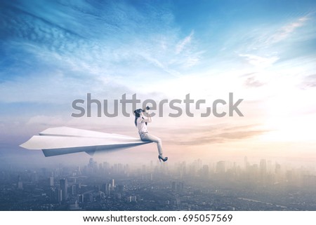 Picture of a female worker sitting on a paper aircraft while flying above a city and looking through a binoculars