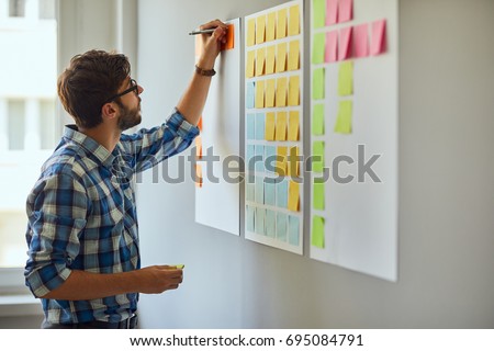 Young creative man writing down ideas on wall full of sticky notes Royalty-Free Stock Photo #695084791