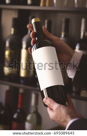 close up of hands of Caucasian men with white shirt holding a red wine bottle with white blank label against the sale shelves of a wine store or restaurant in natural light Royalty-Free Stock Photo #695089645