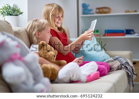 Profile view of joyful little girl showing her sister funny photo on digital tablet while they spending free time together at home