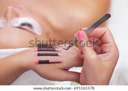 The procedure for eyelash extensions in the beauty salon, eyelashes on the hand of the make-up artist. In the background a girl with long eyelashes.