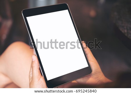 Mockup image of woman's hand holding black tablet pc with blank white screen on thigh in modern cafe