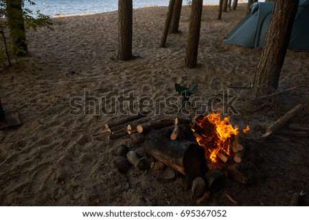 Close up summer outdoor picture of flames and glowing embers in campfire with camping tent in background. Hikers or trekkers spending weekends outdoors by the water, relaxing from busy urban life