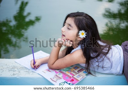 Beautiful small girl lying face down barefoot by the garden pool, smiling, looking to camera and drawing on sketch book with pencil, red bowl with pencils by the pool on the ground, back to school