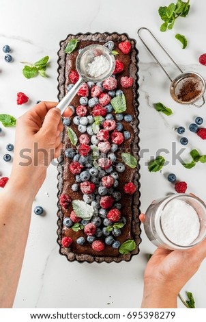 Summer homemade baked pastry. Chocolate cake tart with chocolate cream, blueberry raspberry, mint leaves, woman sprinkle powdered sugar. White marble table, close top view copy space hands in picture
