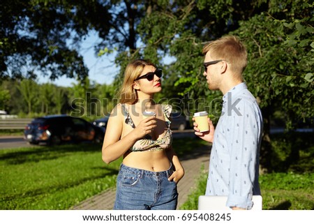 Picture of cute couple drinking coffee outdoors, standing and talking against green trees bakground. Beautiful stylish young man and woman having first date in city park, enjoying coffee-to-go