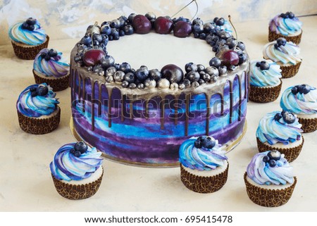 Bright festive blue cake with berries and chocolate and cupcakes with cream .