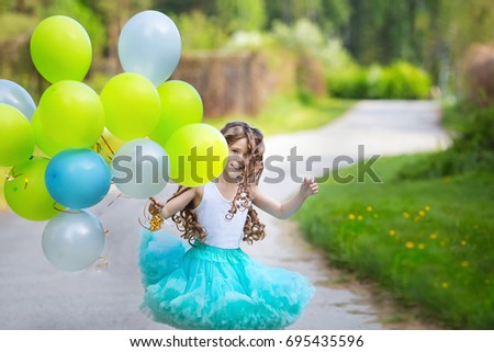  Little beautiful smiling girl with long curly hair wearing smart fluffy skirt playing in the spring garden holding in hands a large bunch of balloons. Happy childhood concept