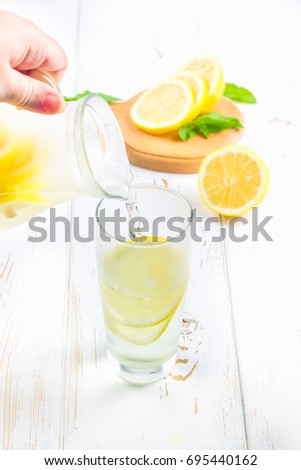 In a glass beaker, a cold lemonade is poured from a jug on a white wooden background surrounded by lemons