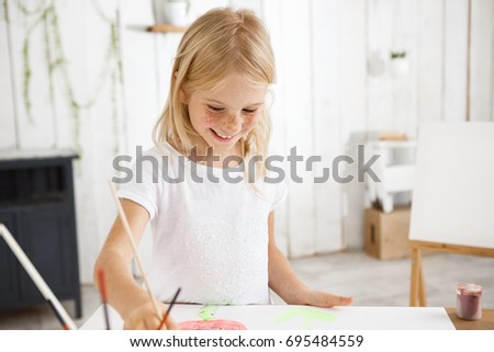 Smiling and cheerful, full of joy child with blonde hair and freckles holding brush in her hand and aspiringly painting picture at the art room. Shot of happy blonde little girl using watercolors
