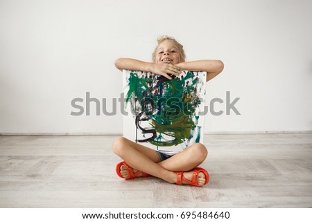 Indoor portrait of blonde smiling with her teeth little girl sitiing cross-legged on the floor, hugging picture she painted for her parents. Happy female child being proud of herself. People and