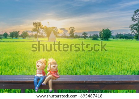 Abstract silhouette soft blurred and soft focus of sunset with the boy and girl cartoons dolly on the wooden seat, green paddy rice field, the beautiful sky, and cloud.By lens flare effect tone.