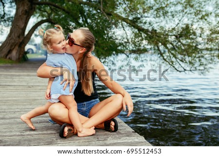 Group portrait of happy white Caucasian mother and daughter child having fun outside. Mom and child girl playing, hugging kissing in park on pier by water lake. Candid authentic real lifestyle.