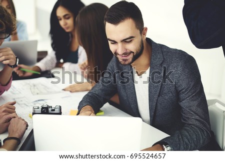 Young bearded employee satisfied with completing successful project using laptop computer connected to wifi sitting with crew of skilled colleagues collaborating on blurred background in office
