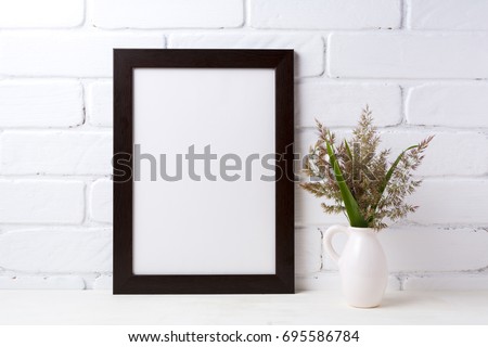 Black brown  frame mockup with meadow grass and green leaves in pitcher vase near painted brick wall. Empty frame mock up for presentation design. Template framing for modern art.