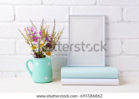 White frame mockup with white chamomile and purple field flowers in mint green pitcher vase and books. Empty frame mock up for presentation artwork. Template framing for modern art.
