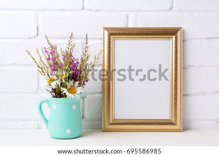 Golden frame mockup with white chamomile and purple field flowers in mint green pitcher vase. Empty frame mock up for presentation artwork. Template framing for modern art.