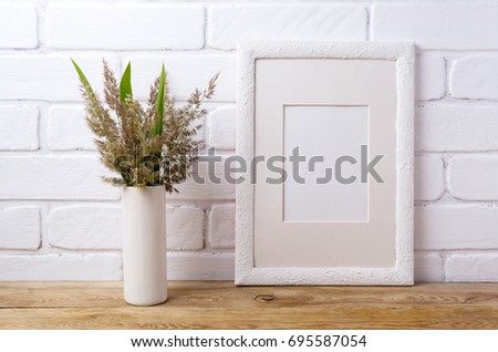 White picture frame with mat  mockup with meadow grass and green leaves in cylinder vase near painted brick wall. Empty frame mock up for presentation artwork. Template framing for modern art.