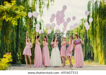 Beautiful happy smiling bride with bridesmaids in light trendy pink dresses on walk outdoors holding pink balloons in green summer city park. Friends, maid of honor, female friendship, wedding concept Royalty-Free Stock Photo #695607151