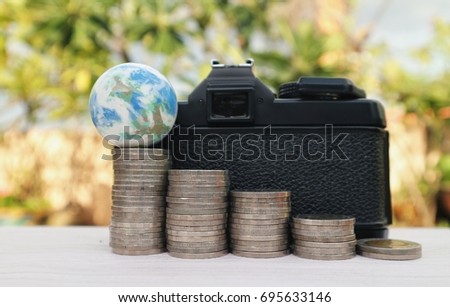 Saving money fro travel concept, world globe on top of rolls stair step coins and black vintage camera on white wooden pattern table, natural garden and bright light background 