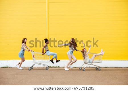 Image of young smiling women friends over yellow wall. Have fun with shopping trolley.