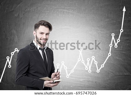 Portrait of a smiling young bearded businessman in a black suit and a tie holding a planner and a pen and looking at the viewer. He is standing near a blackboard with a rising graph