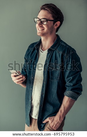 Handsome young man in casual clothes and eyeglasses is using a smart phone and smiling, on gray background