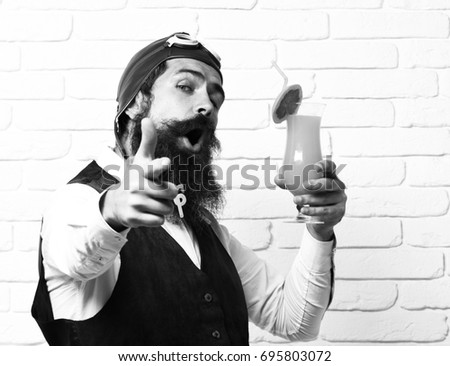 handsome bearded aviator man with long beard and mustache on satisfied face holding glass of alcoholic cocktail in vintage suede leather waistcoat with hat and glasses on white brick wall background