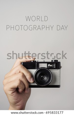 closeup of the hand of a young man with a retro film camera and the text world photography day against an off-white background