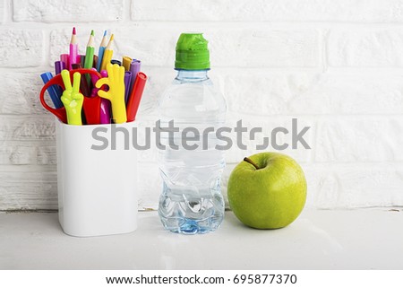 Against a white brick wall, a schoolchild's table for homework: a glass with pens and pencils, a bottle of water and a fresh apple for a snack. Selective focus