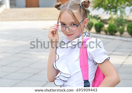 Smiling cute schoolgirl of primary school in glasses. Girl with a pink bag near the building of the school outdoors.