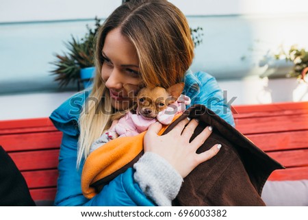 Beautiful young woman sitting on bench and holding adorable little Chihuahua dressed in winter jacket. 