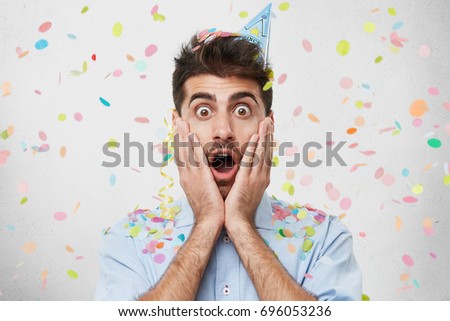 Shocked man in holiday cap and shirt, keeping his hands on cheeks, looking with bugged eyes and opened mouth into camera, celebrating his birthday, being surprised to see huge present from friend