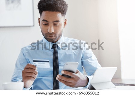 Picture of confident young dark-skinned office worker with Afro hairstyle holding credit card in one hand and mobile phone in other, paying for flowers he sent to his wife online via internet