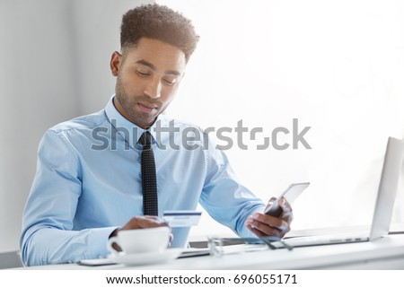 Picture of attractive young African American manager dressed in formal wear sitting at his workspace with cell phone, looking at information on his credit card while signing up for mobile banking app