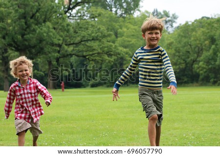 Two young boys are running together in the park. One child is dressed in red and white plaid and the other has a stripped shirt. They are playing outdoors