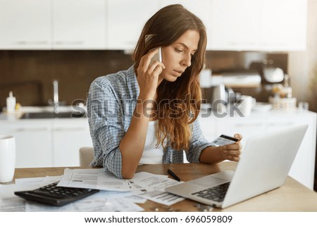 Beautiful young Caucasian woman calling bank using cell phone concerning information on credit card that she is holding. Serious female connecting to mobile banking service using electronic device
