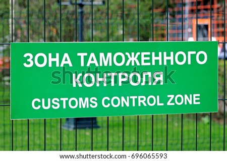 Green sign with the inscription in Russian and English: Customs control zone on the fence of metal rods. Side view