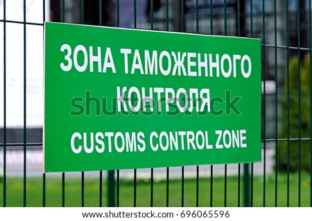 Green sign with the inscription in Russian and English: Customs control zone on the fence of metal rods. Side view. Shallow depth of field
