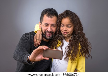 A black father taking selfies with a smartphone while his young daughter hugging him around his neck
