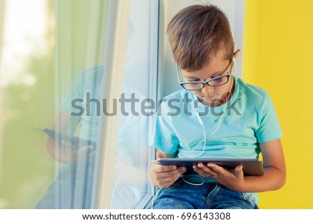 Little boy sits on the windowsill and listening to music through headphones. Child uses the tablet, watching cartoon, playing games, surfing internet.