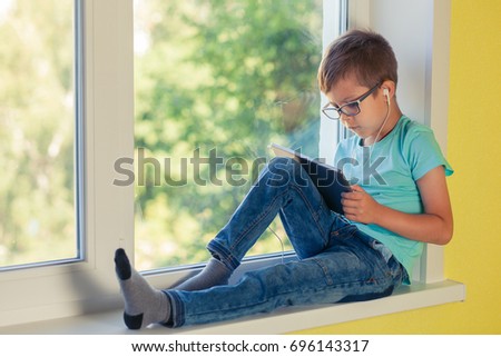 Little boy sits on the windowsill and carefully watching a movie on the tablet. The child is intently watching what is happening on the screen.