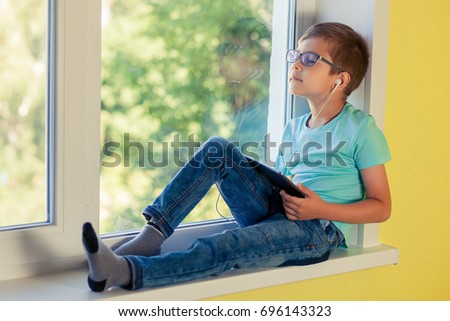 Little boy sits on the windowsill and enjoy listening to music through headphones. Best playlists and new singles. The child uses the tablet to watch cartoon, play games, engage in Internet surfing.