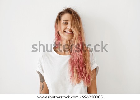Horizontal portrait of pleasant-looking Caucasian female with long hair, pink on tips, having tattooes on arms, wearing white casual T-shirt, covering her face with hair, looking happily in camera Royalty-Free Stock Photo #696230005