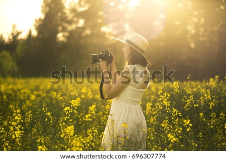 Portrait of young and attractive woman in white dress and with hat in rape field. Soft focus. She is taking a picture of something or somebody.