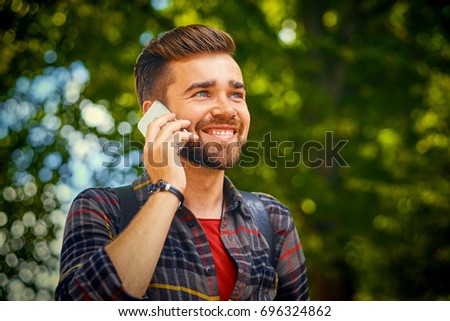 Bearded male dressed in a fleece shirt talks by a smart phone in a park.