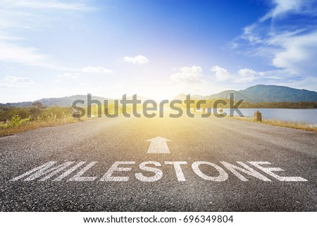 Asphalt road with arrow guideline and Milestone letters painted on the surface. An image of a road milestones are representative of success in the future goal. Road to success with light of the sun Royalty-Free Stock Photo #696349804